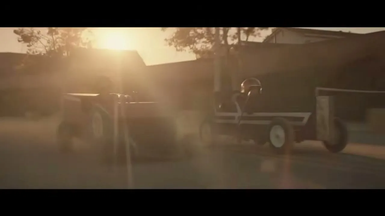 Two children in homemade soapbox derby cars race down a dusty suburban street at golden hour, kicking up dust as they speed past houses.