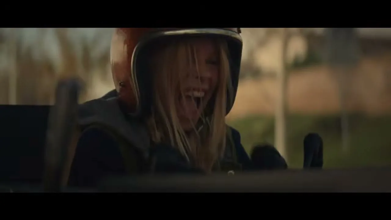 A young girl with blonde hair and a helmet laughs joyfully while riding in a go-kart outdoors during the day.