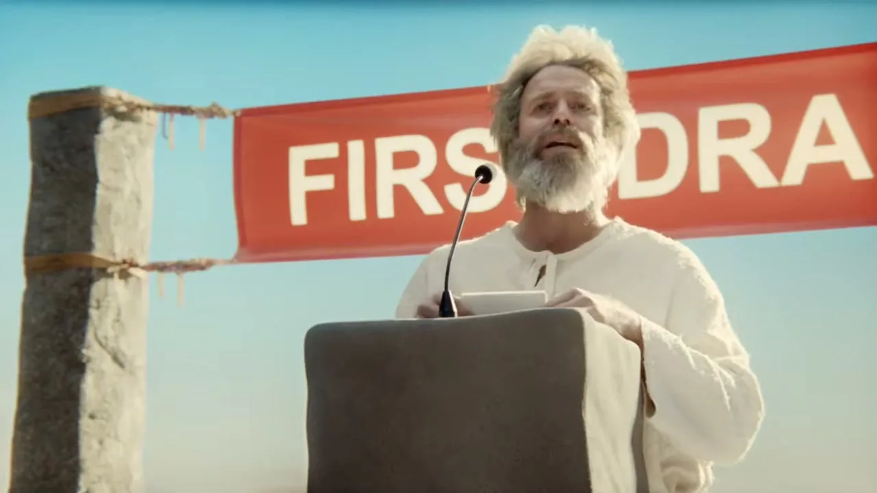A man with a white beard and hair, dressed in white, stands at a podium with a microphone, looking up, with a red banner behind him that reads "FIRST DRA".