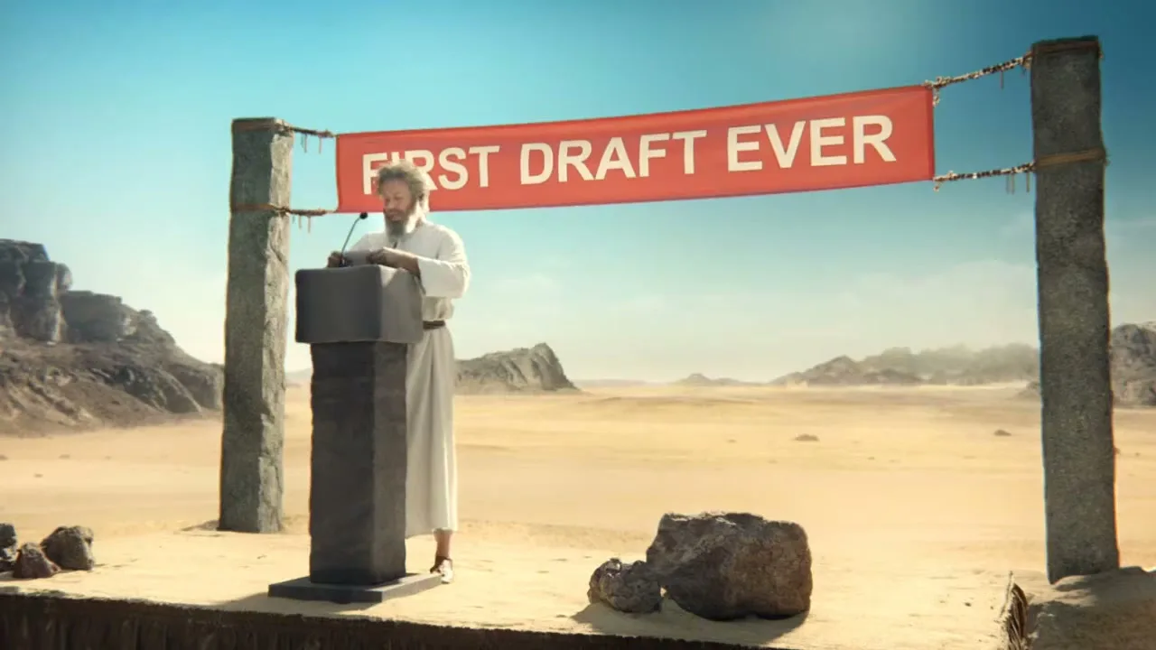 A man with a long beard and white robe stands at a podium in a vast desert landscape under a bright blue sky, with a red banner above him.