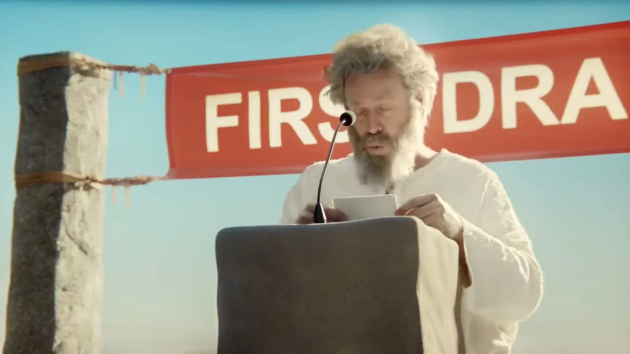 A man with a long beard and white robes stands at a podium, reading from a paper with a red banner behind him that says "FIRST DRA".