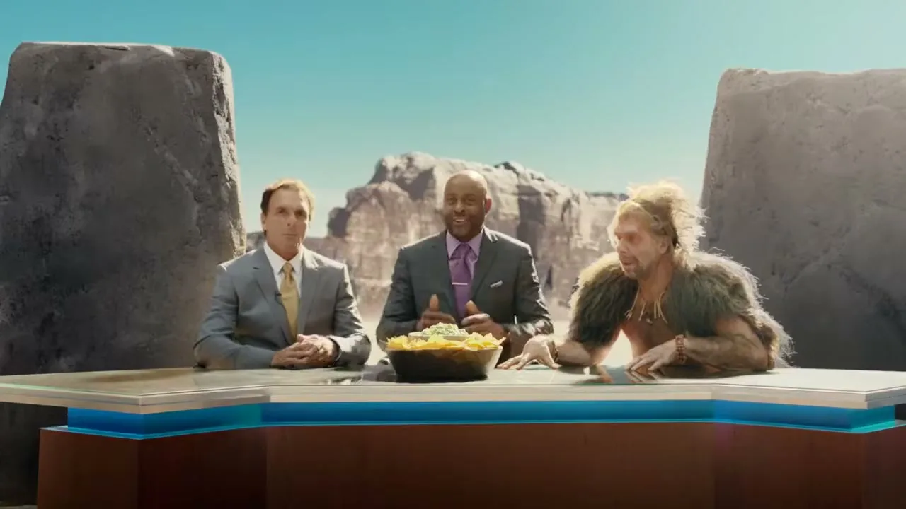 Three men, two in suits and one in caveman attire, sit at a news desk with a bowl of guacamole and chips, discussing avocados in a rocky, outdoor setting.