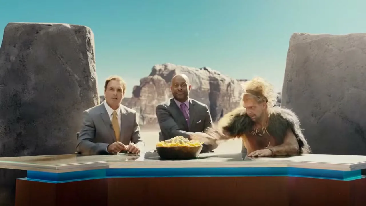 Three men, two in suits and one in caveman attire, sit behind a news desk with a bowl of guacamole and chips, in a desert landscape.