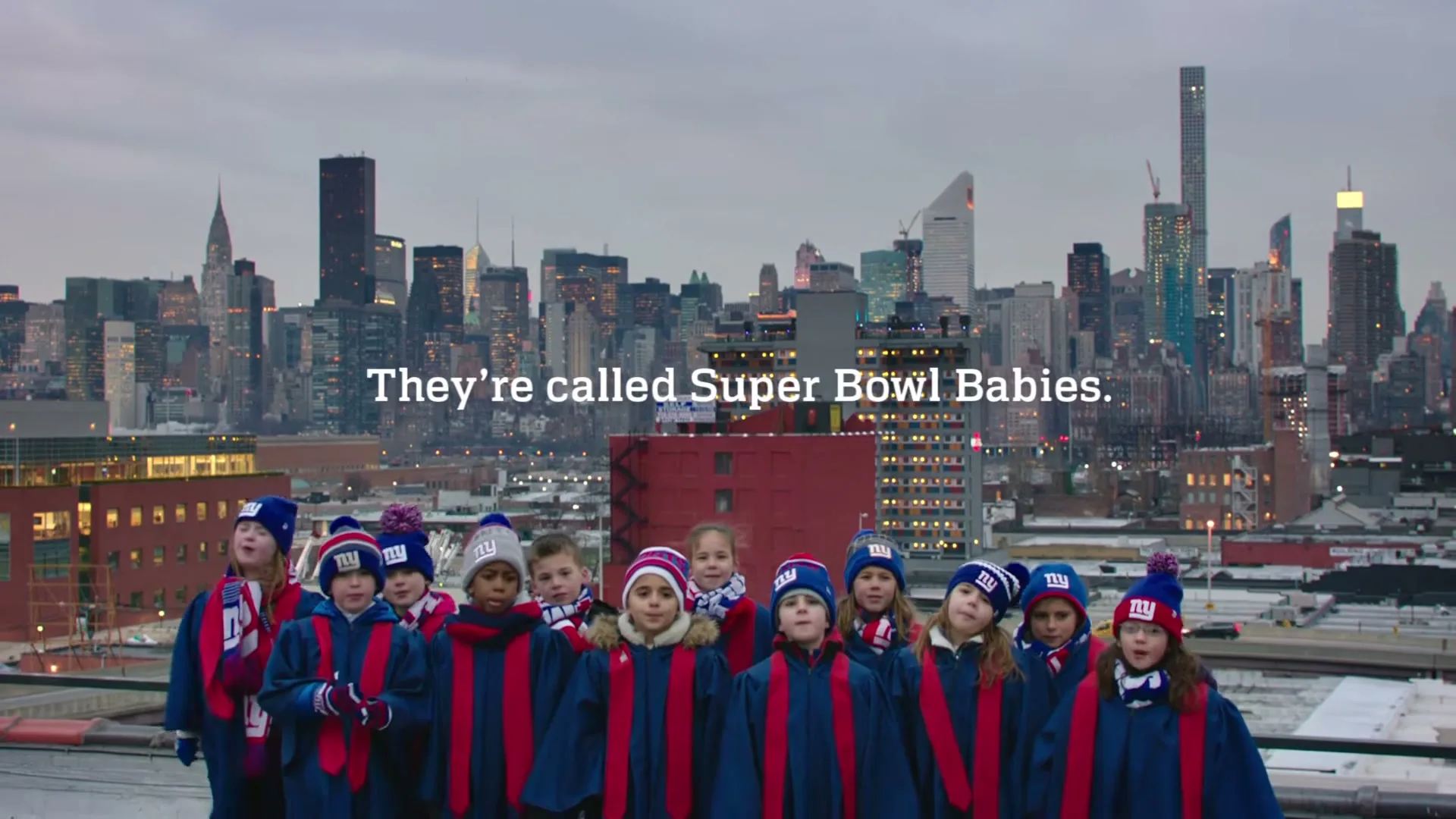 A group of children wearing blue robes and knit hats stand on a rooftop, singing with a city skyline in the background.