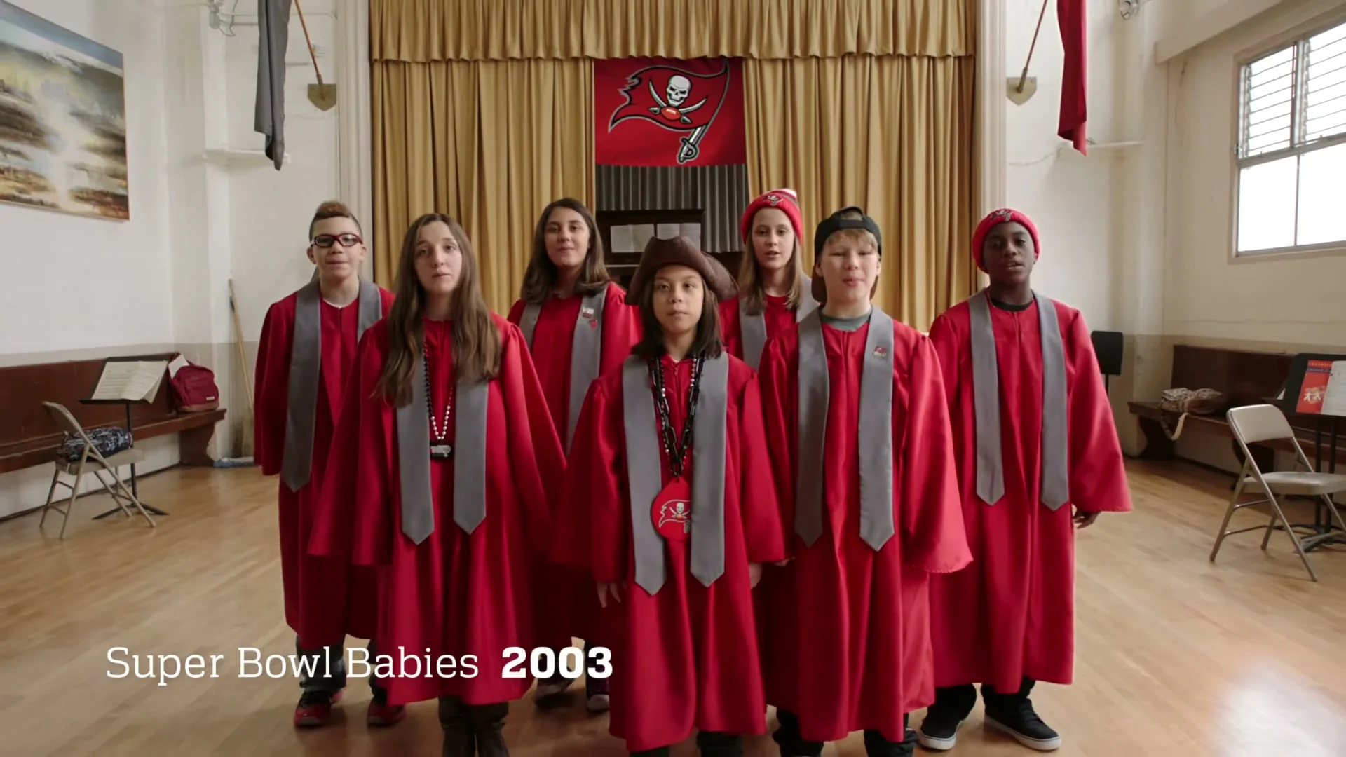 A diverse group of children in red choir robes with gray stoles sing in a school auditorium, with a Tampa Bay Buccaneers flag hanging behind them.
