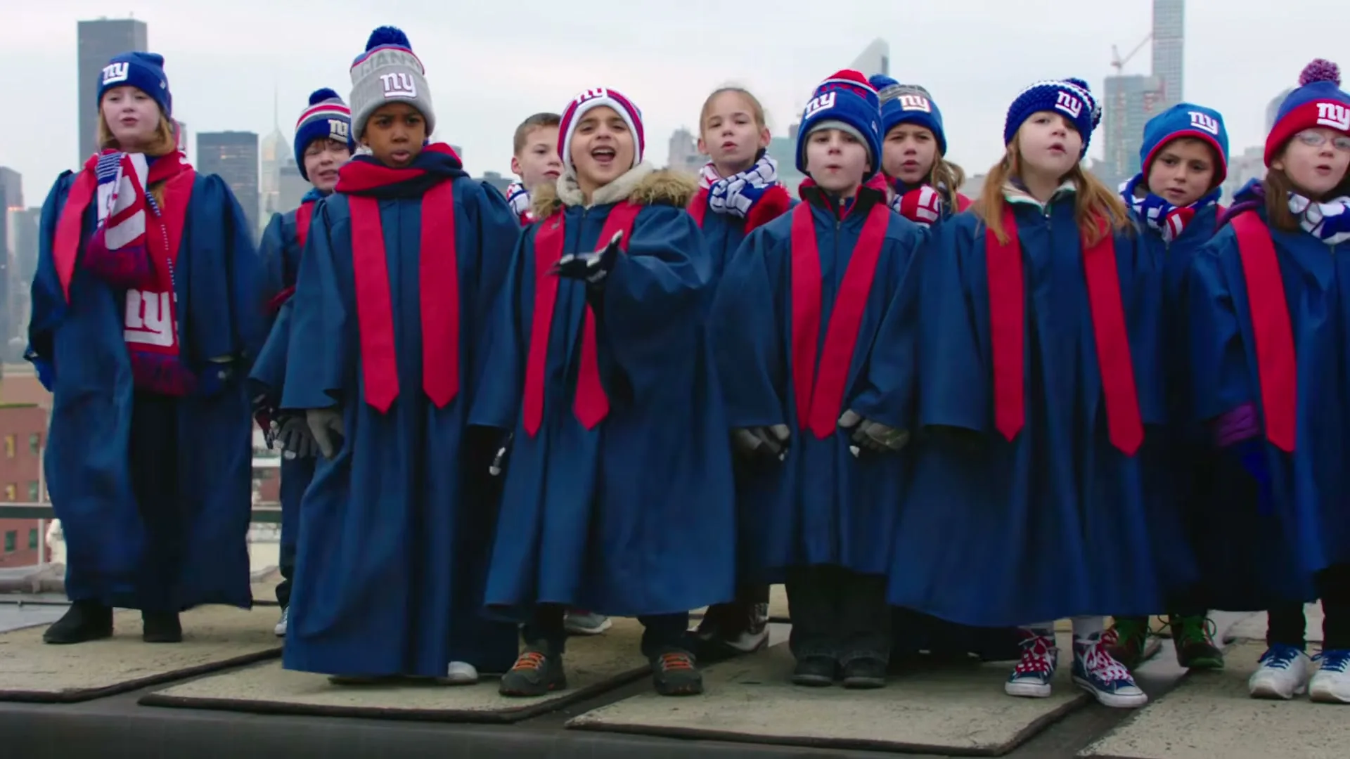 A diverse group of children, dressed in blue choir robes, red stoles, and New York Giants beanies and scarves, sing on a rooftop with a city skyline in the background.