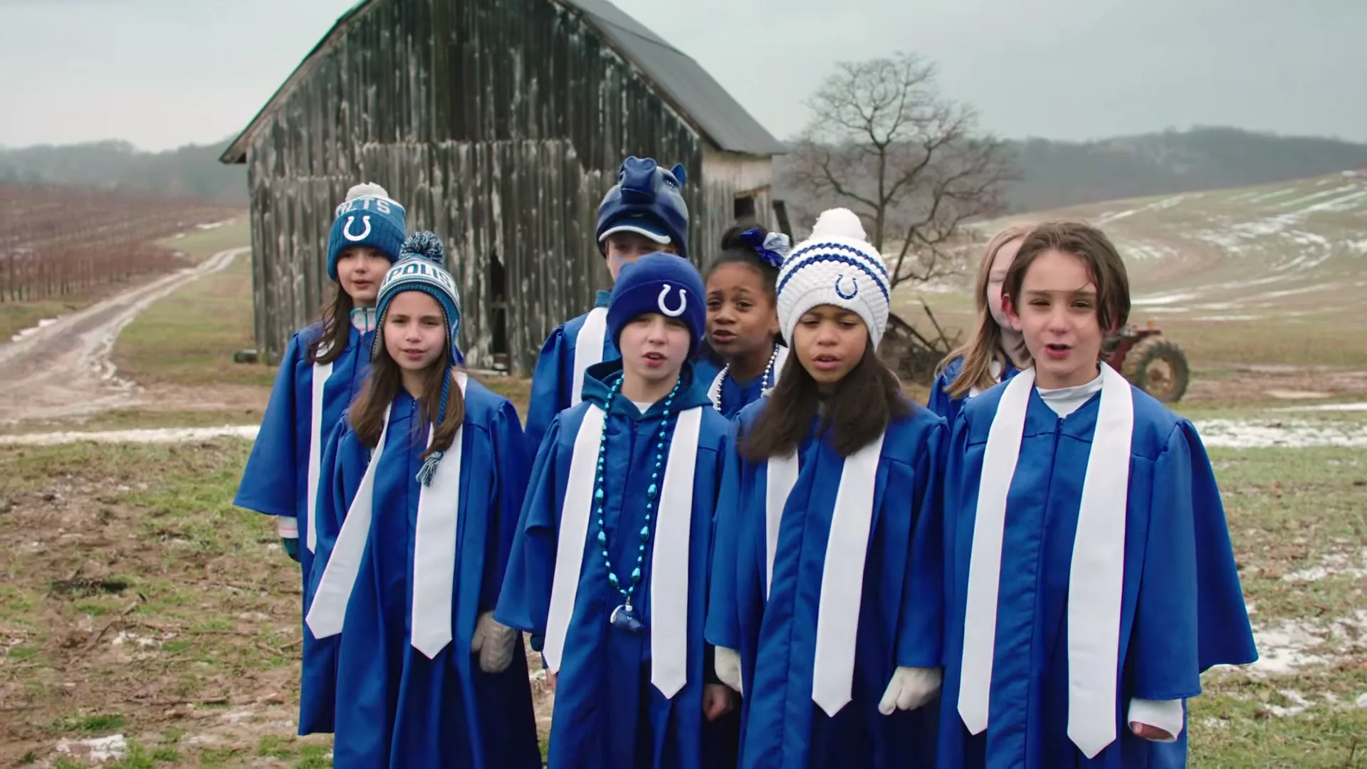 A group of diverse children, dressed in blue choir robes and Indianapolis Colts beanies, sing together in a rural, snowy field with a rustic barn in the background.