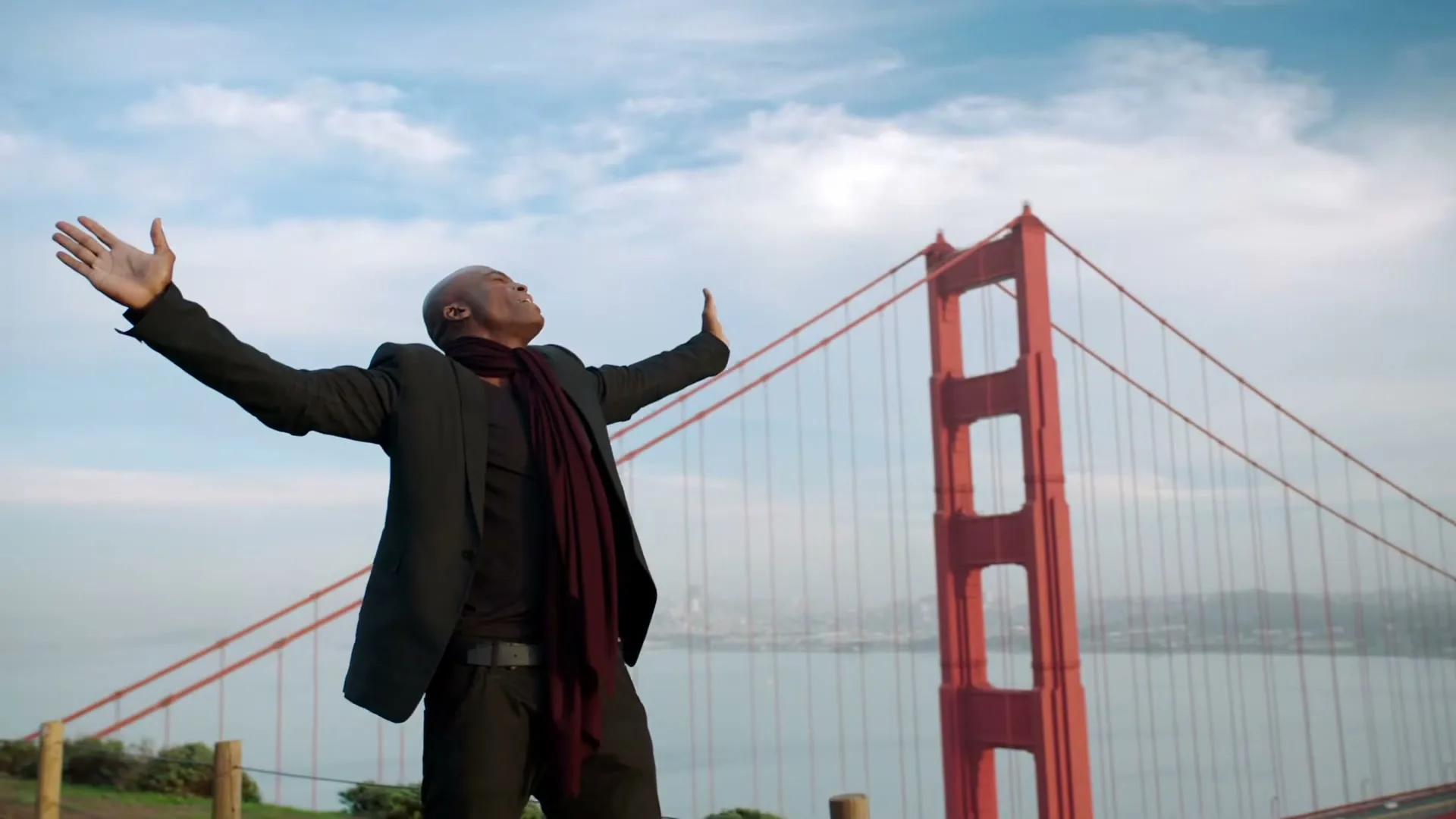 A man with dark skin, wearing a black suit and a red scarf, stands with arms outstretched, looking up at the sky, with the Golden Gate Bridge in the background.