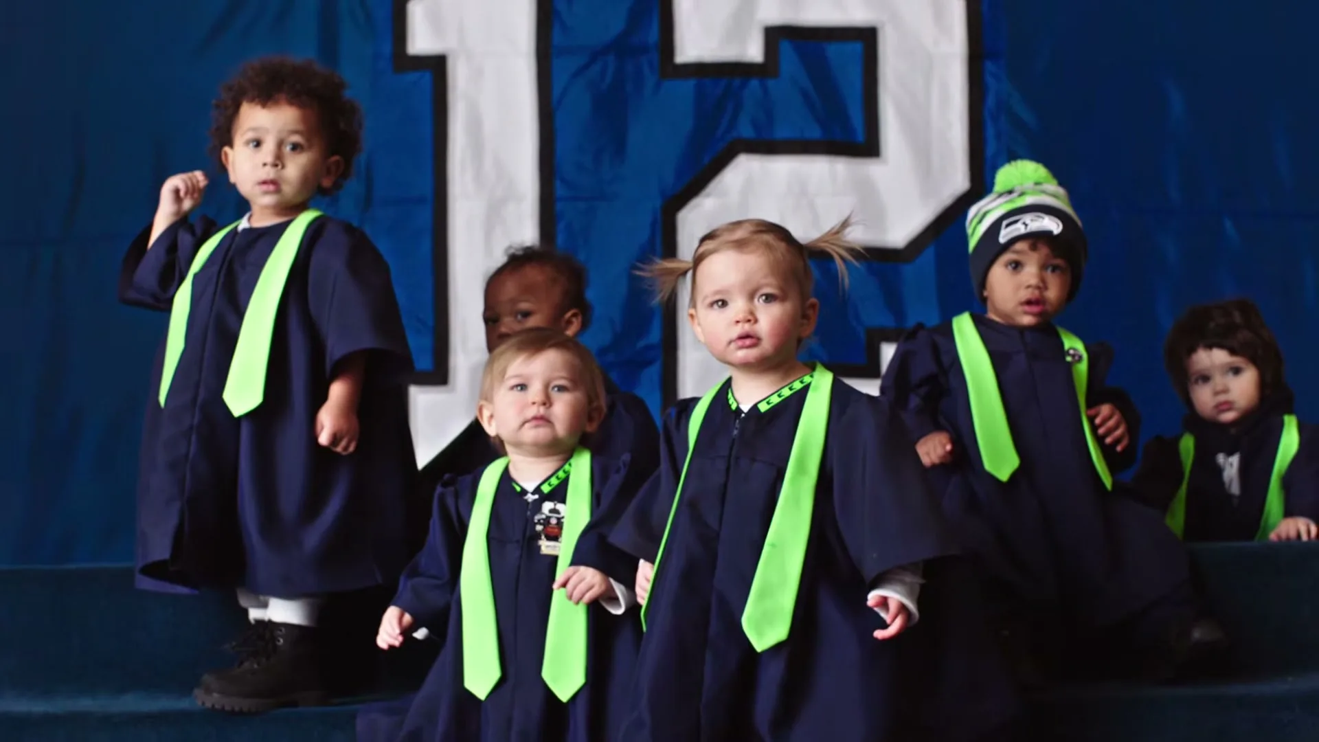A group of diverse babies, dressed in navy blue graduation gowns with bright green ties, stand on risers in front of a large blue banner with a white "12" on it.