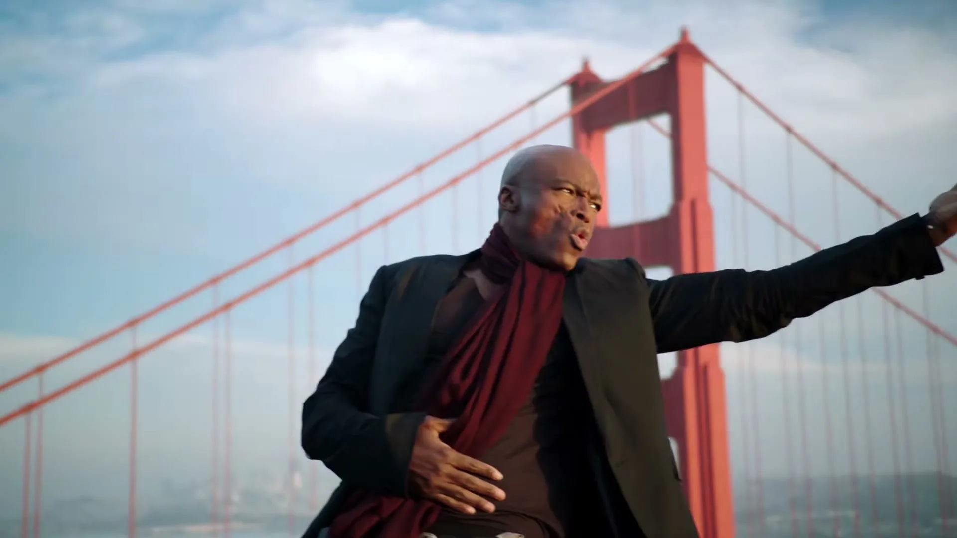 A bald Black man with facial scars, wearing a black suit and a red scarf, sings passionately with outstretched arms in front of the Golden Gate Bridge.