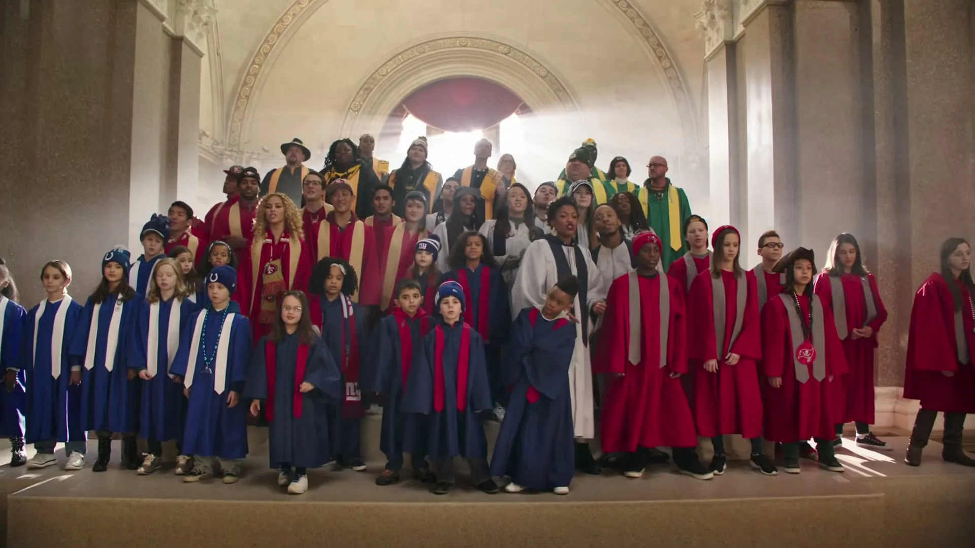 A large choir of adults and children, dressed in red and blue robes, sing together in a grand, sunlit hall with arched doorways.