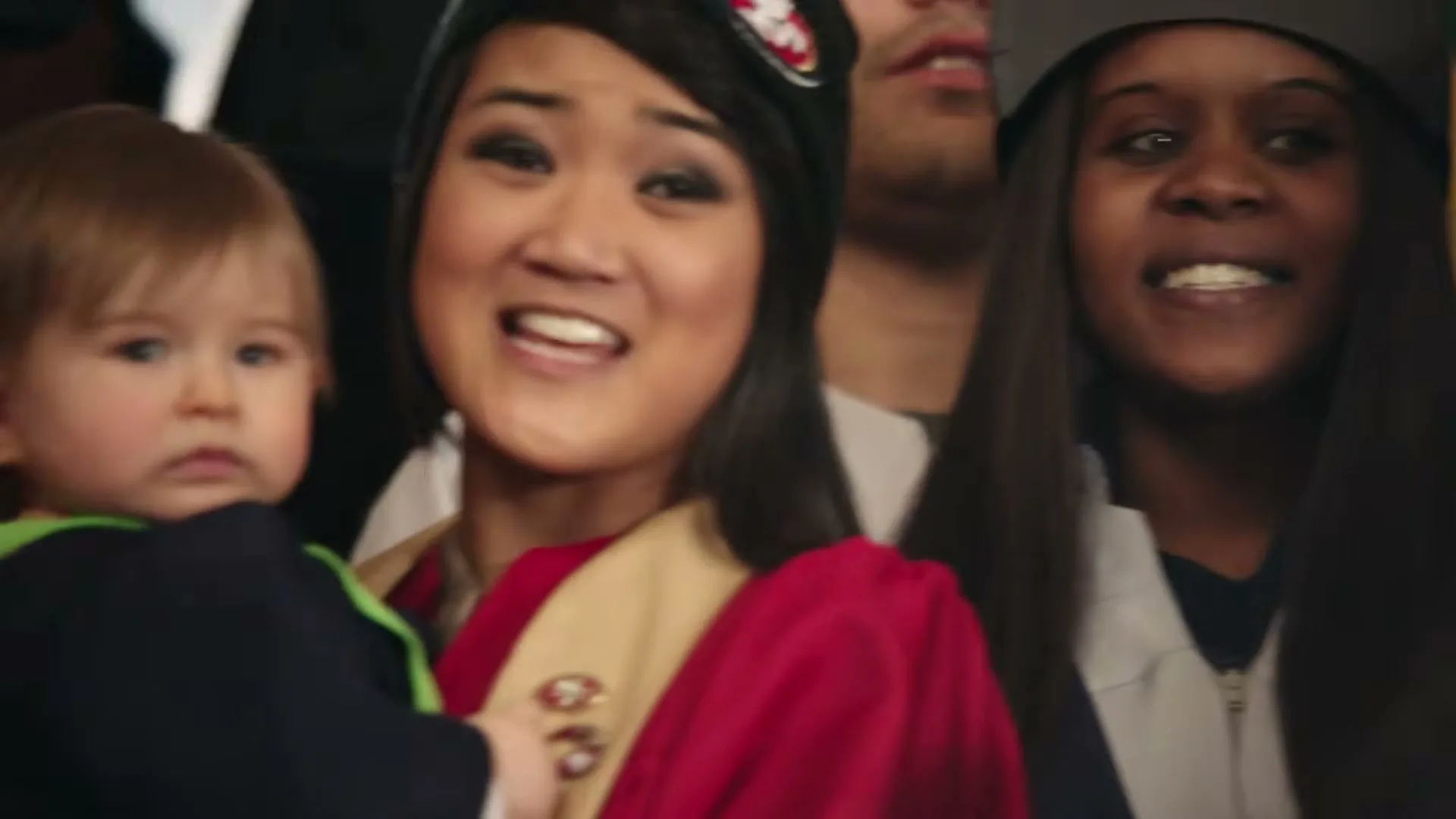 A diverse group of adults and babies, dressed in choir robes and NFL team gear, sing together in a close-up shot.