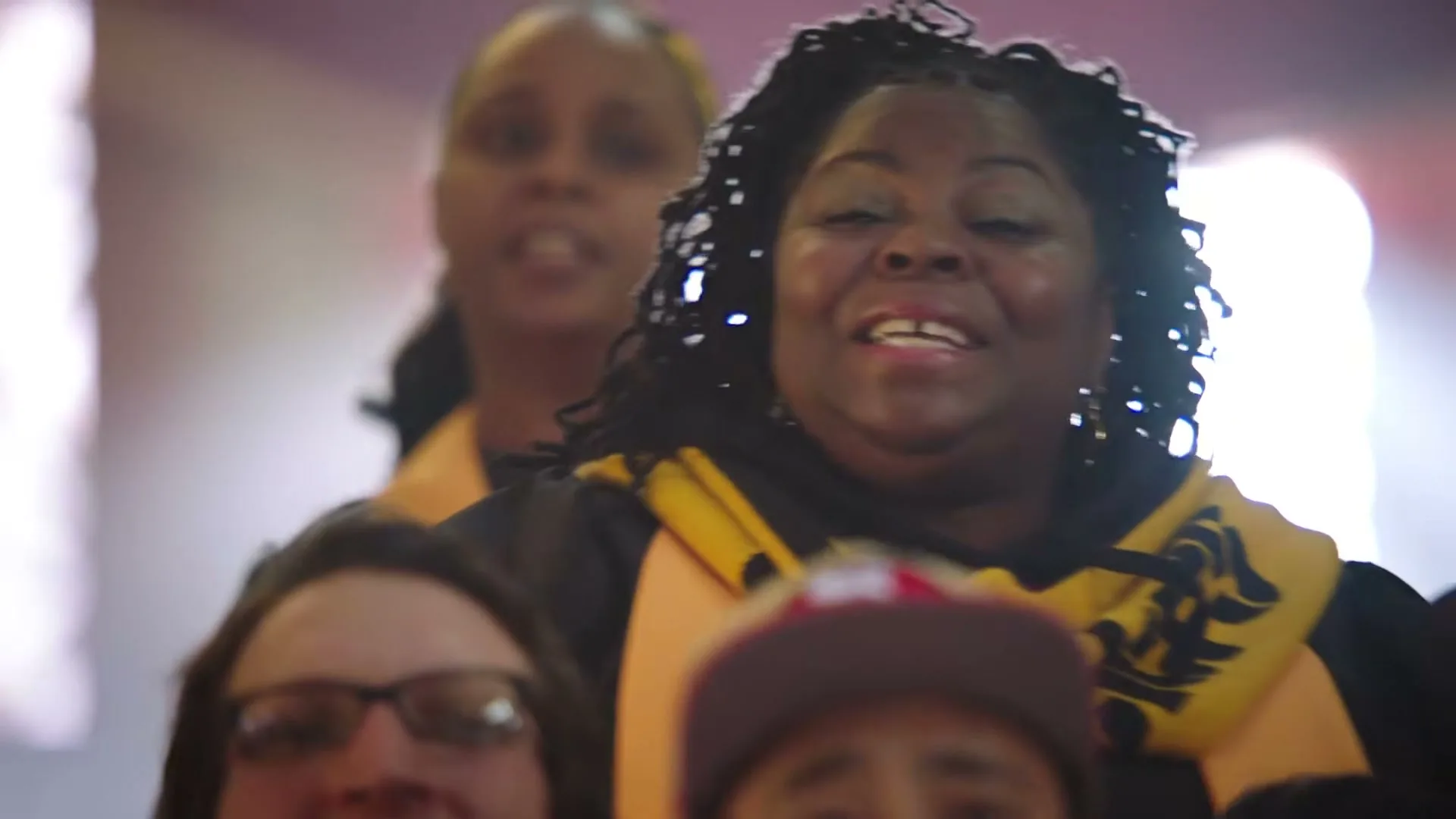 A Black woman with dreadlocks, wearing a black shirt and yellow scarf, sings joyfully in a choir at a stadium.