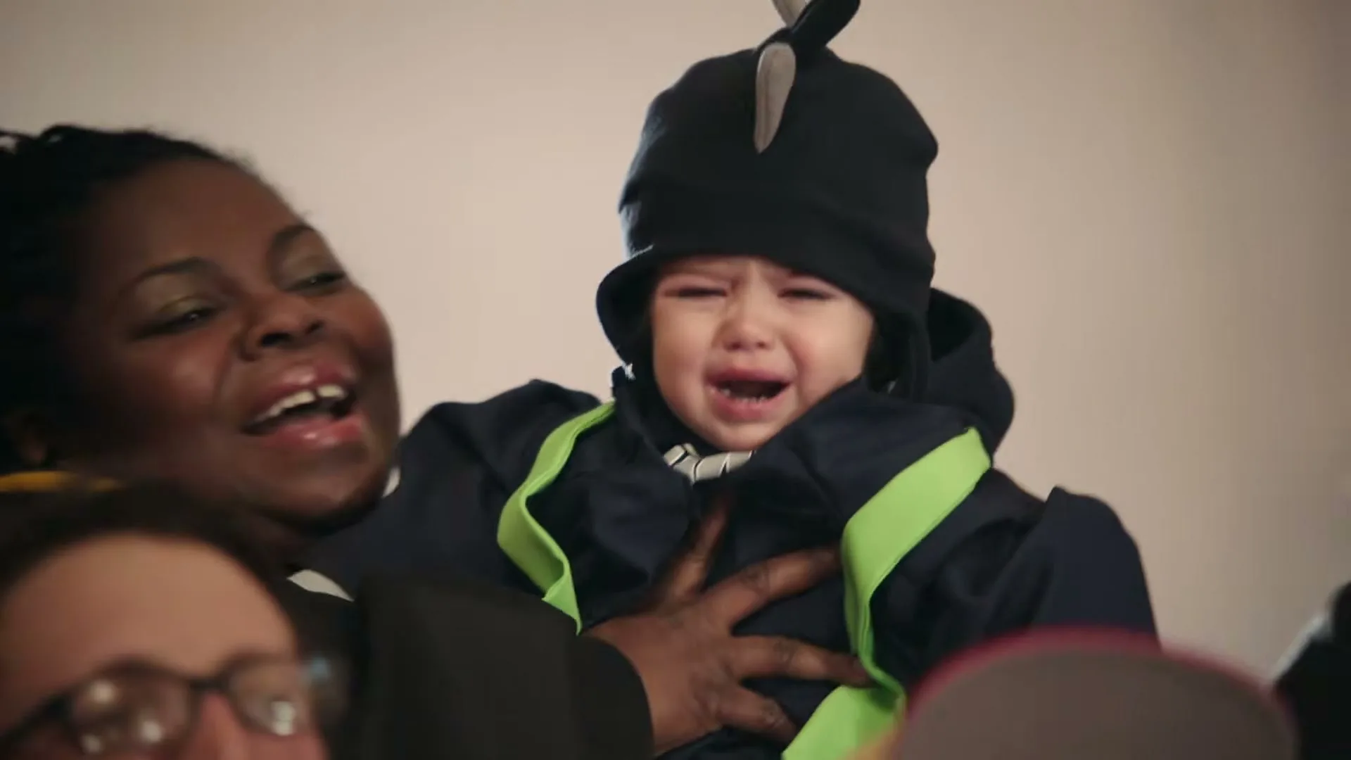 A Black woman in a black and yellow top holds a crying baby dressed in a blue and green outfit and a black hat, surrounded by other people.