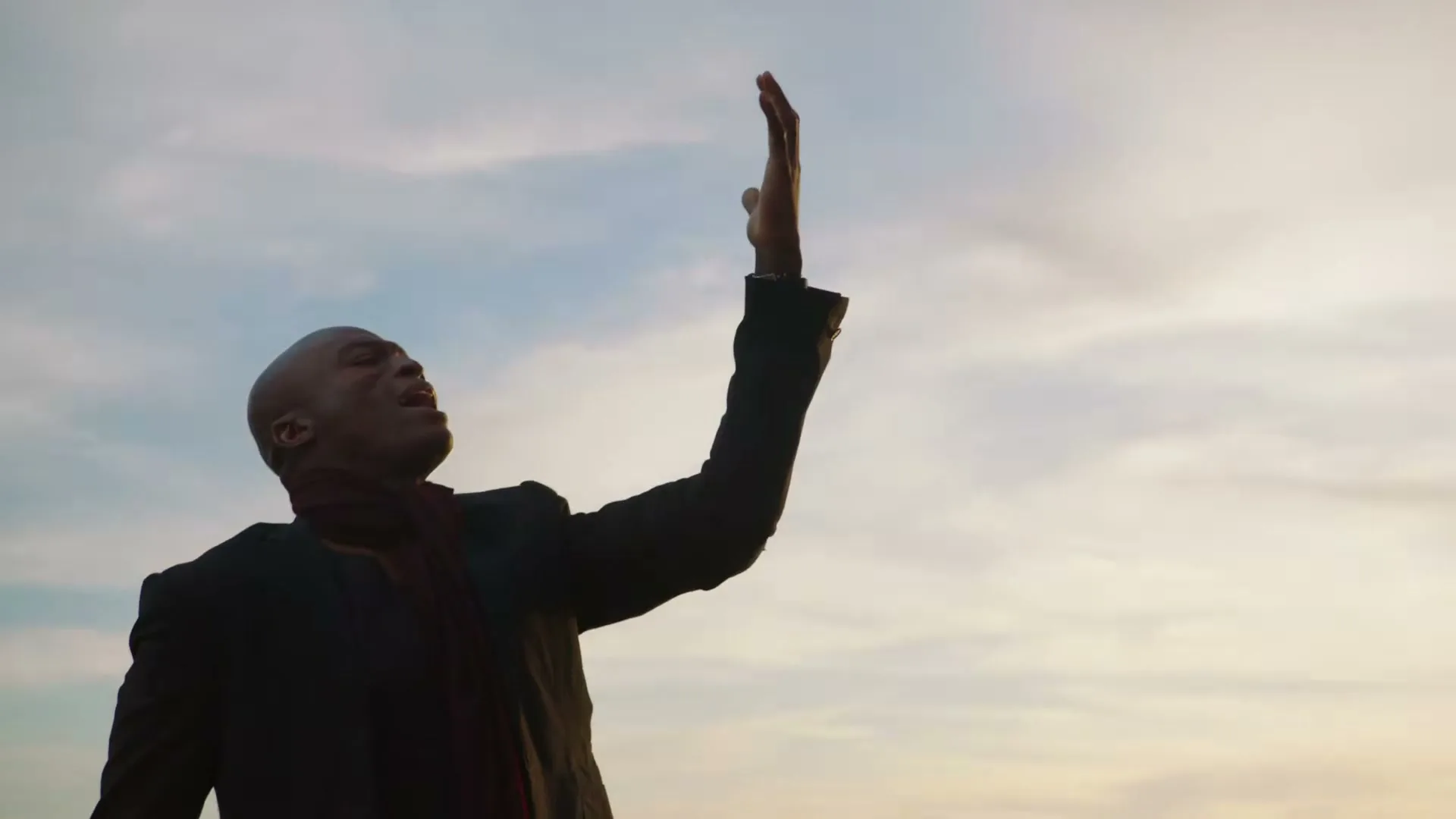 A bald African American man in a suit and scarf sings with his arm raised, looking up at the sky during golden hour.
