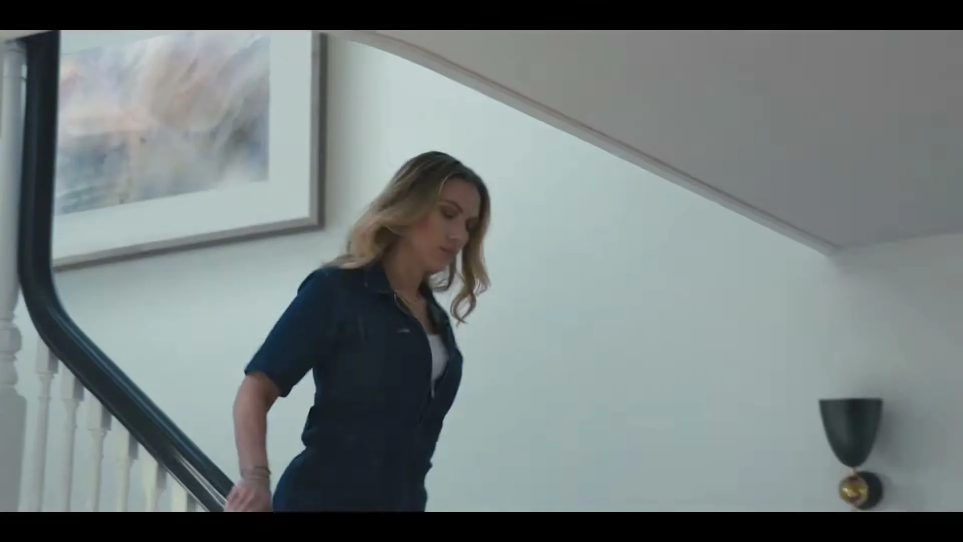 A woman with blonde hair, wearing a dark blue jumpsuit, walks down a white staircase in a modern home.