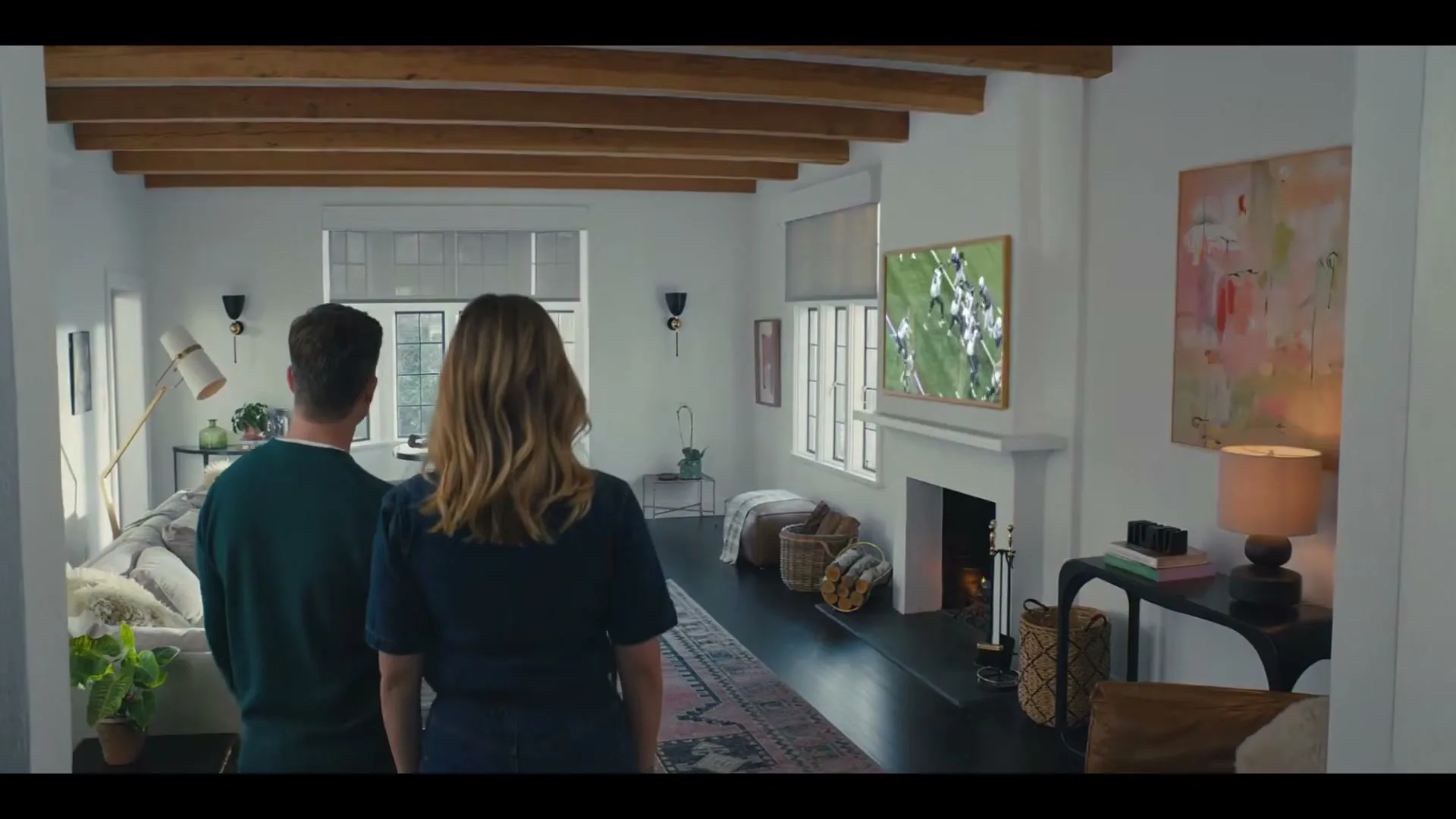 A couple stands in their living room, watching football on a large TV screen mounted above a fireplace, as the blinds automatically close.