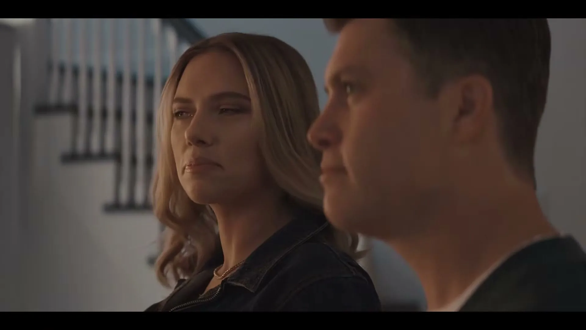 A woman with blonde hair and a man with brown hair look forward, with the woman appearing slightly annoyed, in a modern home.