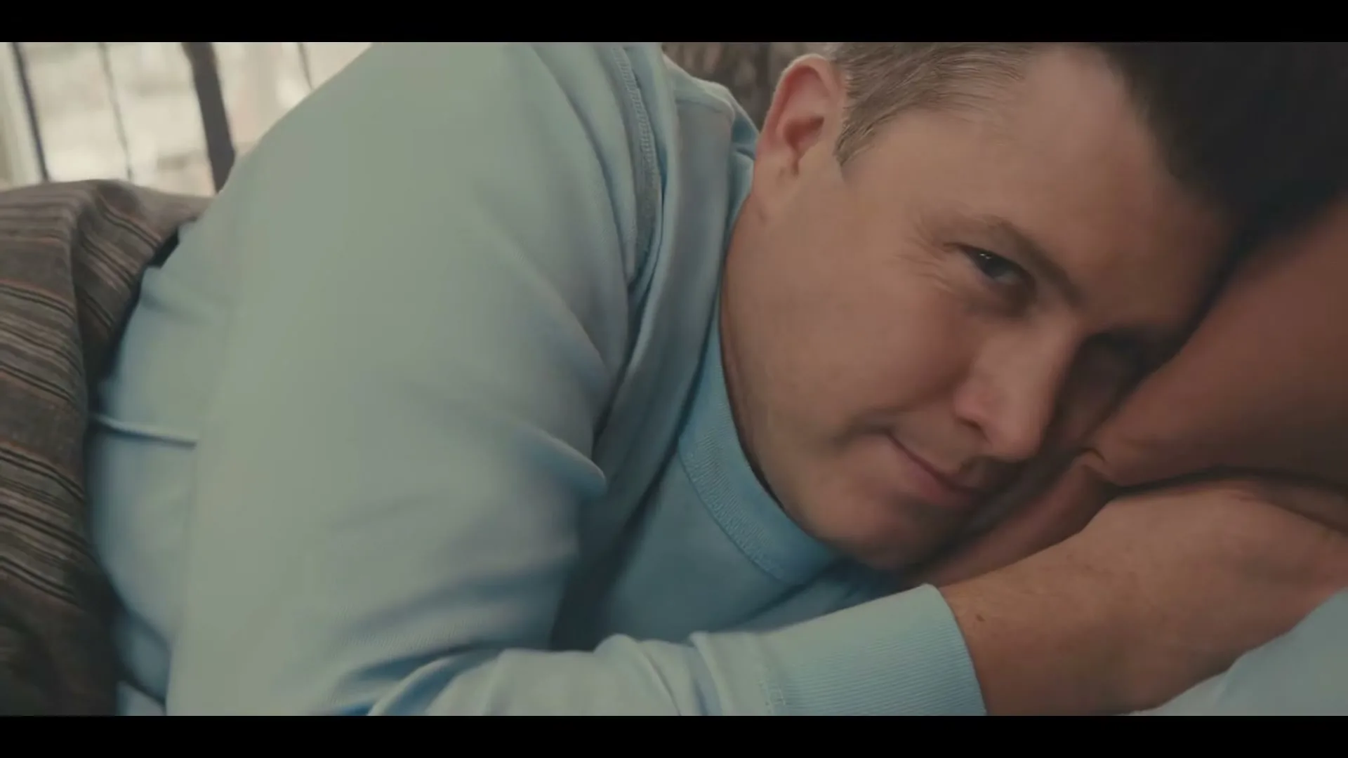 A man in his 30s, wearing a light blue long-sleeved shirt, lies in bed, smiling and then closing his eyes, in a bedroom.
