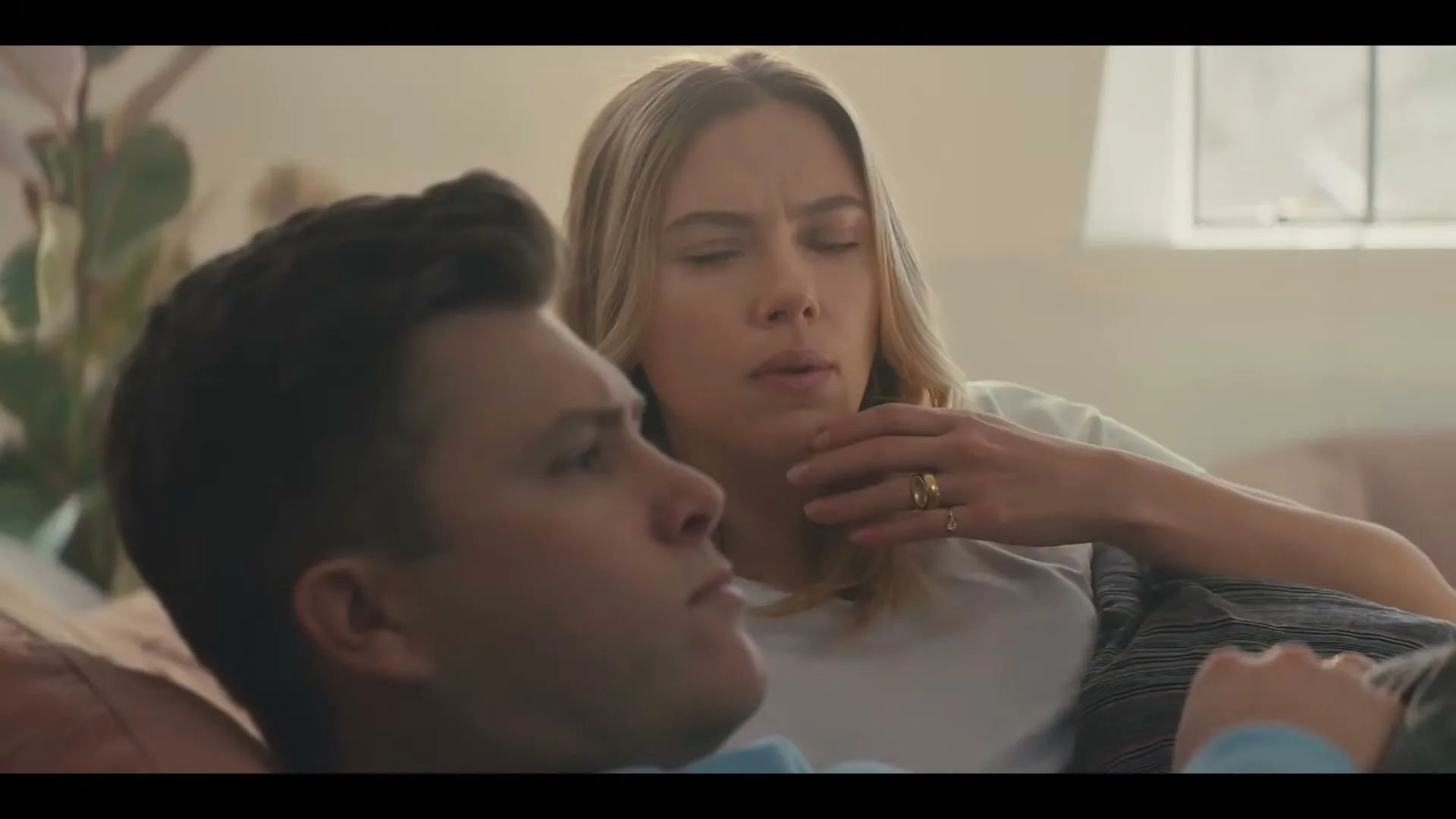 A woman with blonde hair and a white t-shirt looks thoughtfully at a man lying on a couch, who appears bored.