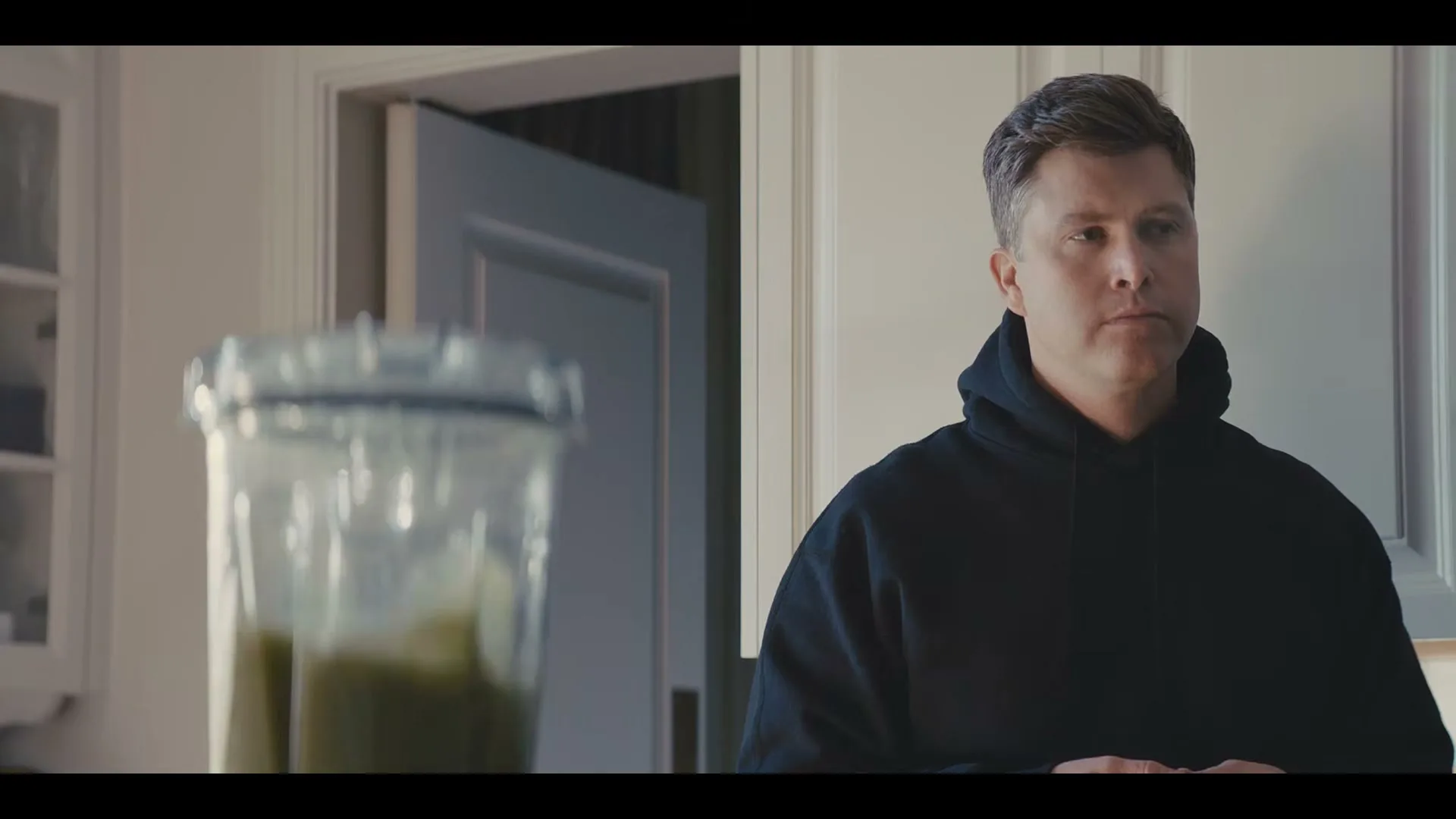 A man in a black hoodie stands in a kitchen, looking at a blender with a green smoothie.