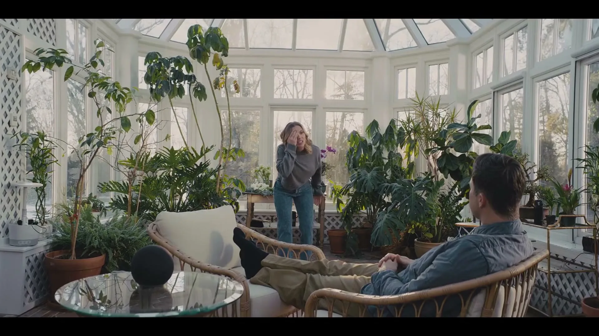 A woman in a grey sweater and jeans stands frustrated in a sunroom full of plants, while a man relaxes in a chair, looking at her.