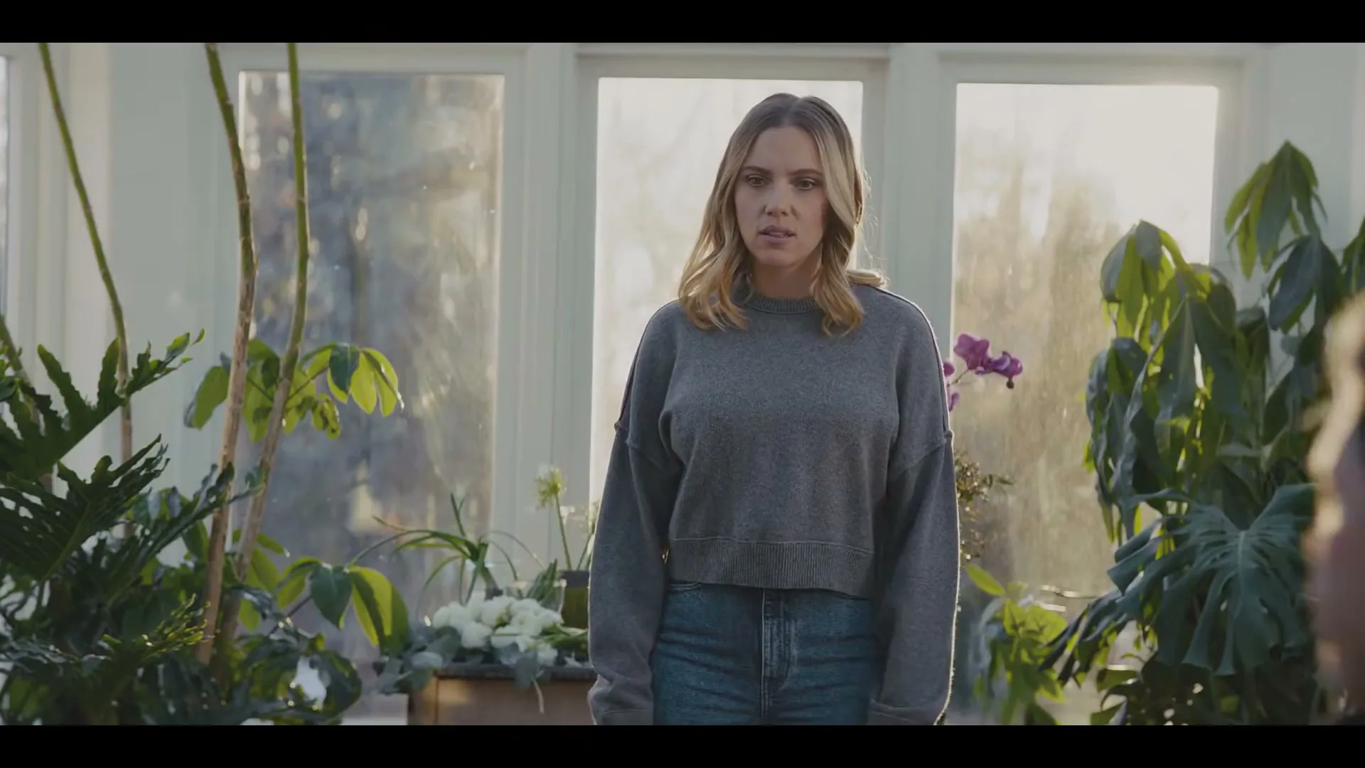 A woman with blonde hair and a grey sweater looks confused and surprised in a sunlit room filled with plants, as a man's head is visible in the foreground.