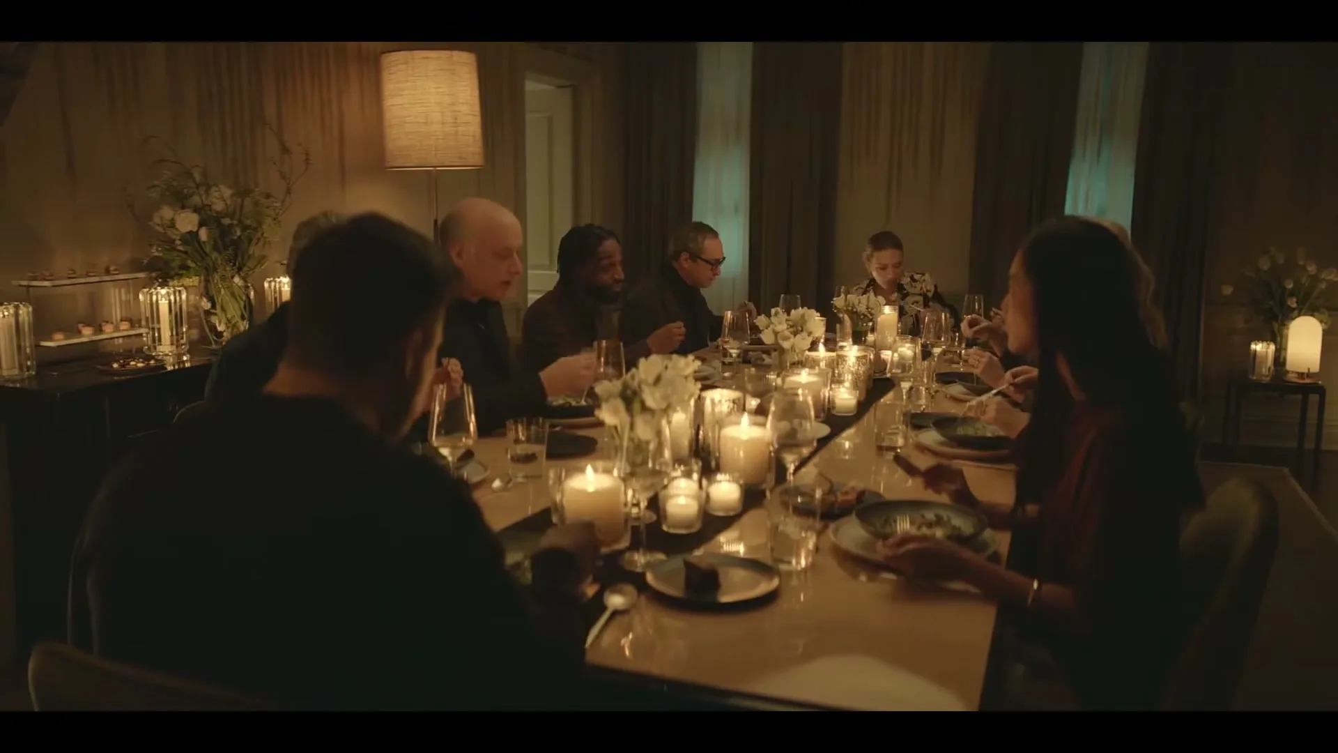 A group of people are seated around a long dining table, eating dinner by candlelight in a dimly lit room.