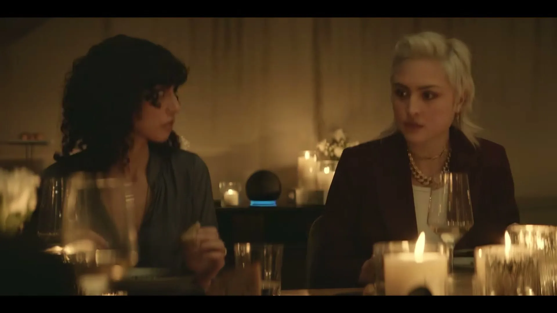 Two women with dark and blonde hair sit at a candlelit dinner table, looking at each other as an Amazon Echo glows blue in the background.