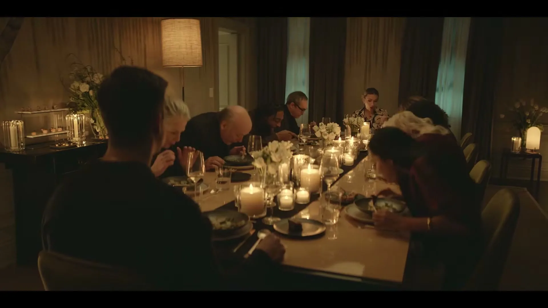 A group of people, including a man in a black shirt, are seated around a candlelit dining table, eating in a dimly lit room.