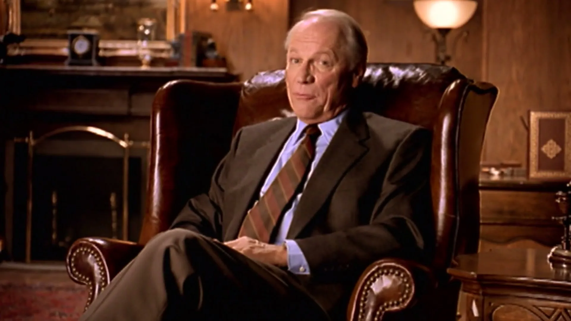 A senior man in a suit sits in a leather armchair, speaking directly to the camera in a dimly lit, traditional study.