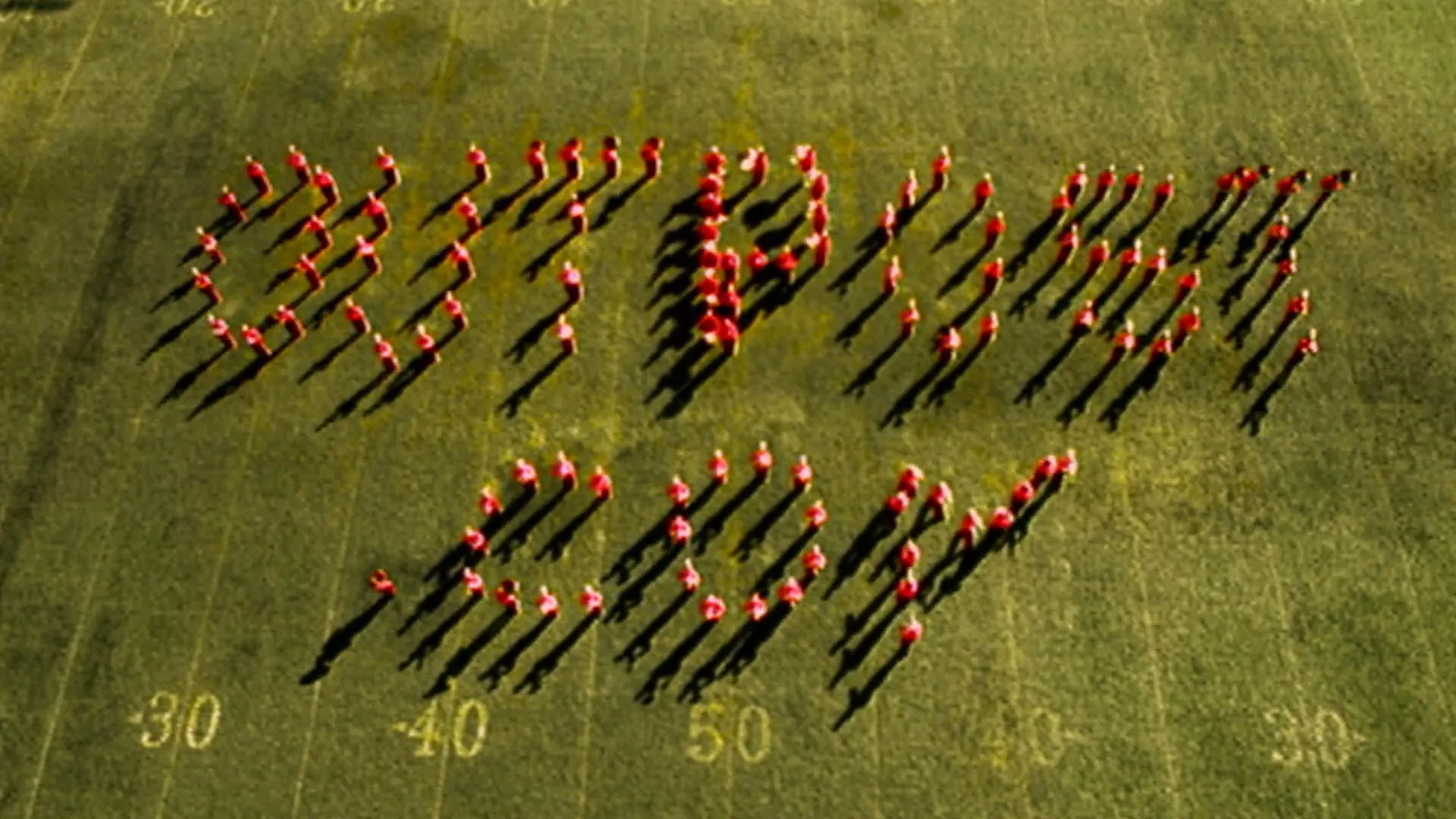 A large group of people in red shirts form the words "OUTPOST.COM" on a green football field, casting long shadows in the afternoon sun.