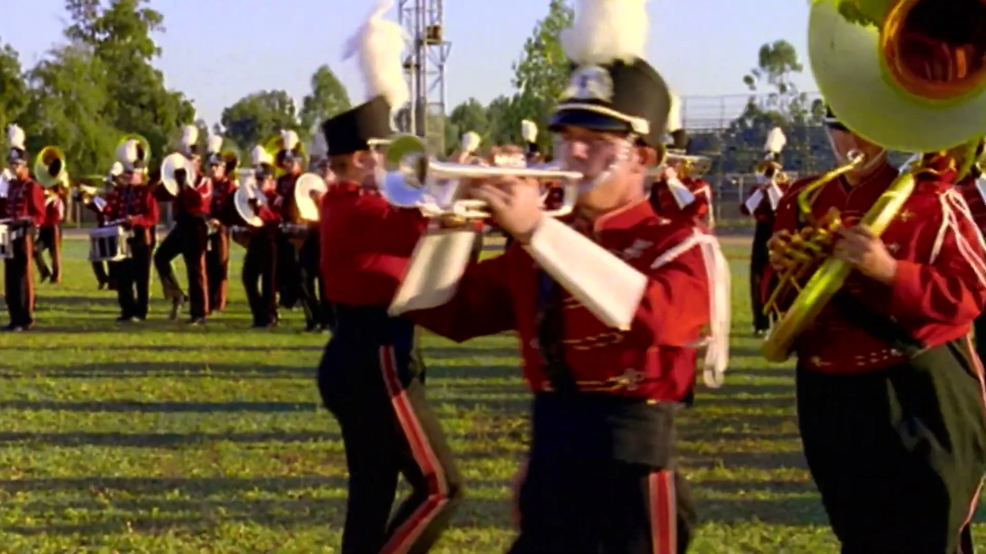 A marching band in red and black uniforms plays instruments and marches across a grassy field during the day.