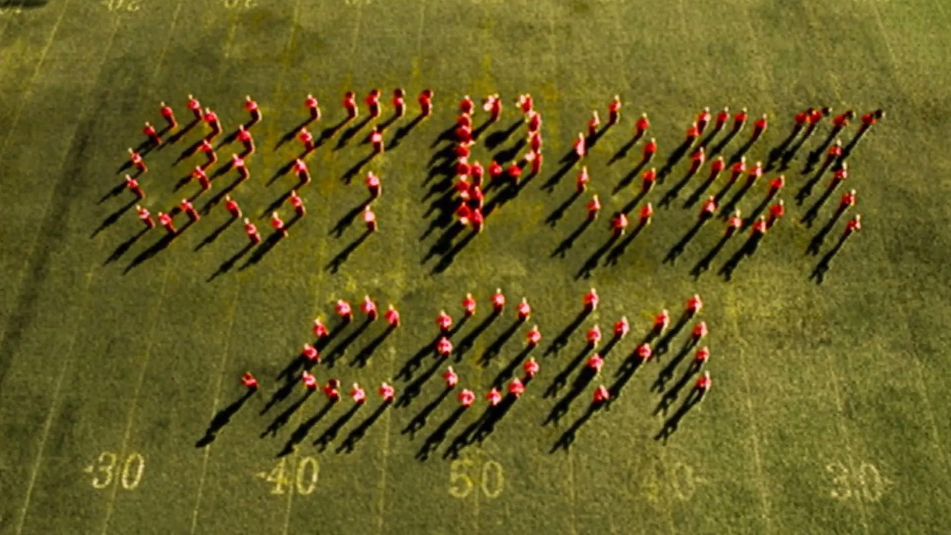 A marching band in red uniforms forms the word "OUTPOST.COM" on a green football field under a bright sky.