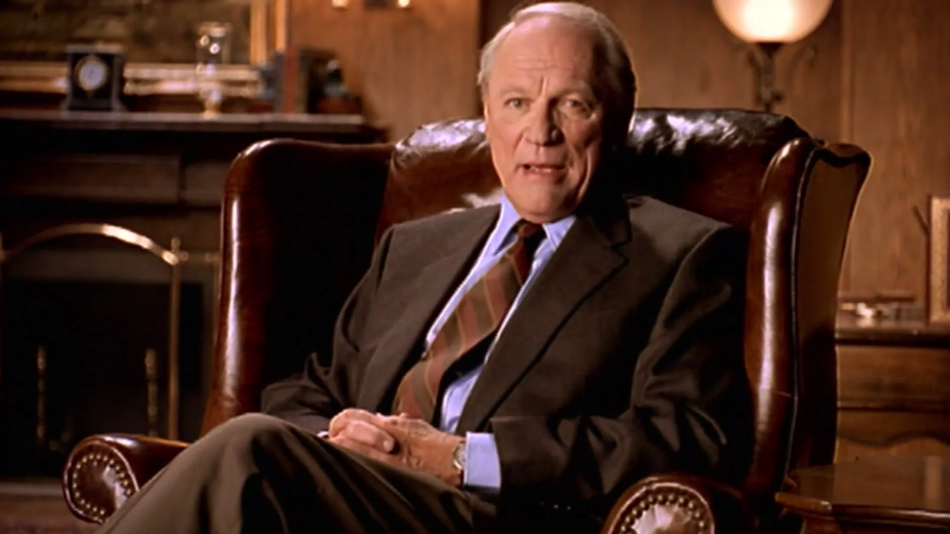 An older man in a suit sits in a leather armchair, speaking directly to the camera in a dimly lit, opulent room.