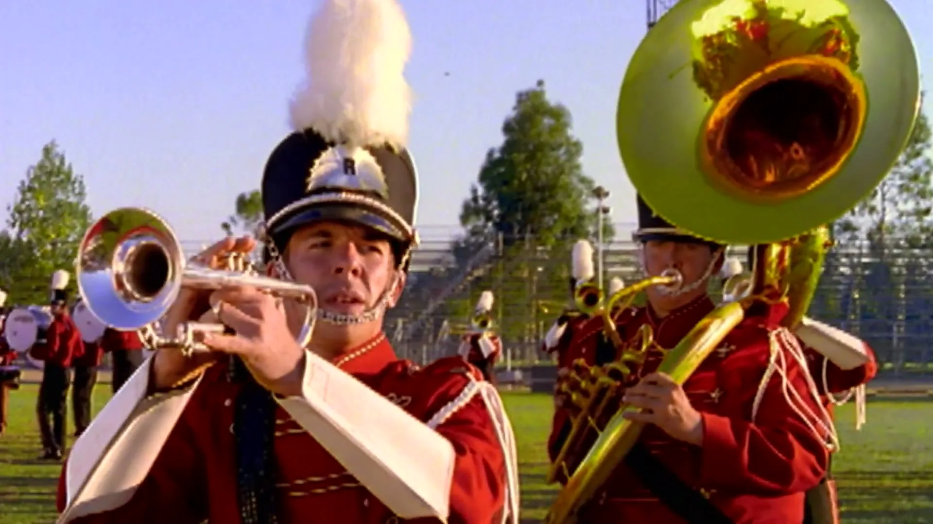 A marching band plays instruments on a sunny day in a stadium, with a trumpet player and a tuba player in the foreground.
