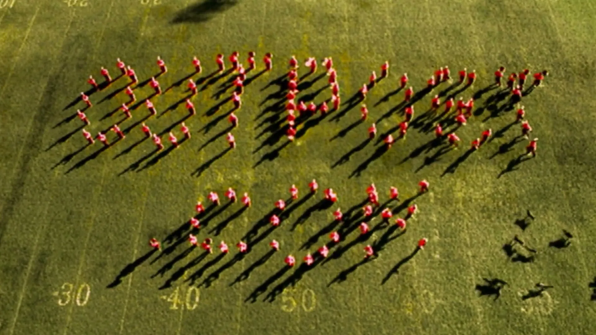 An aerial shot shows a marching band forming the word "DUMMIES" on a green football field under bright sunlight.