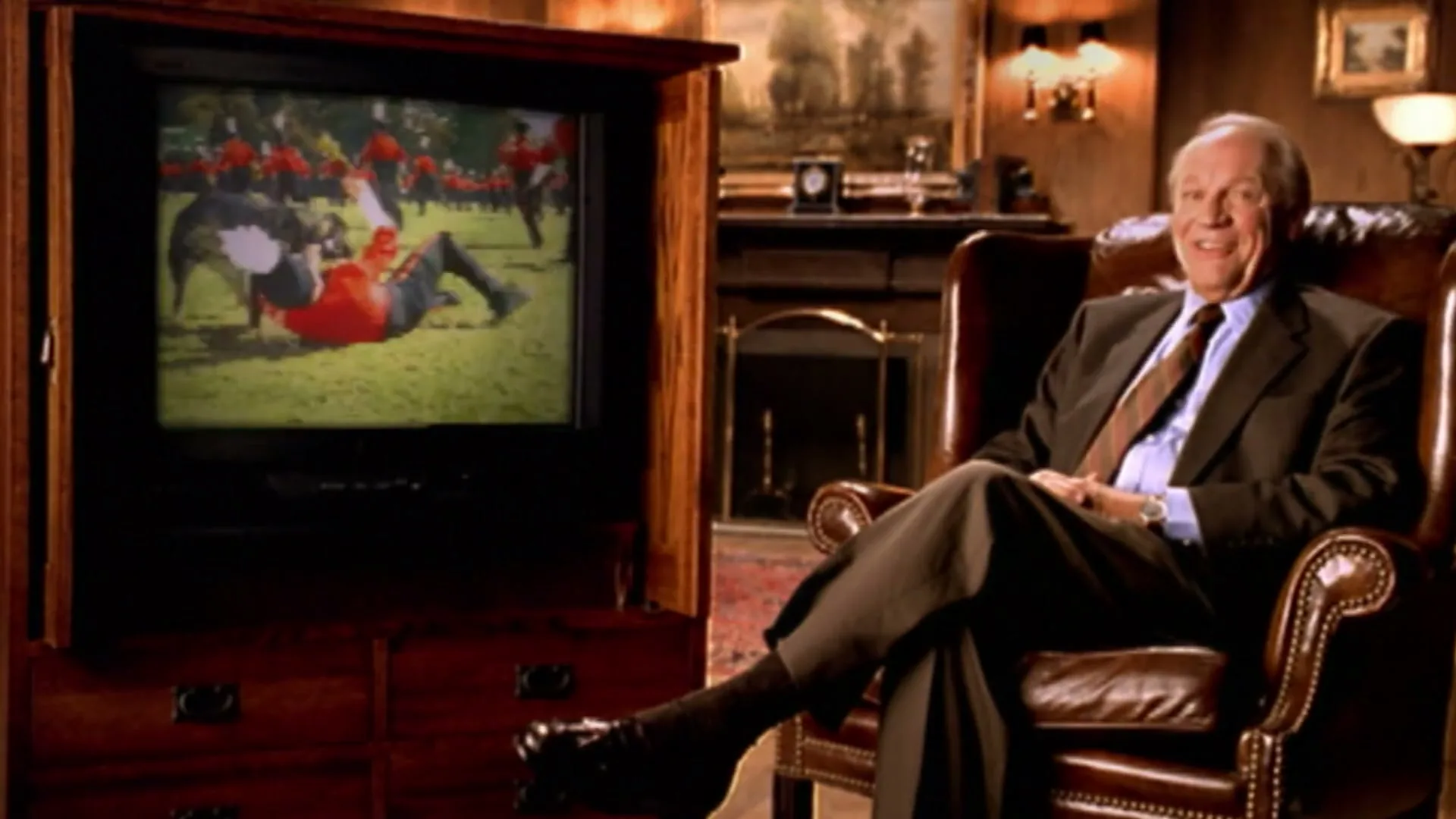 An older man in a suit sits in a leather armchair, smiling and laughing, as he watches a dog attack a marching band member on a TV screen in a wood-paneled room.