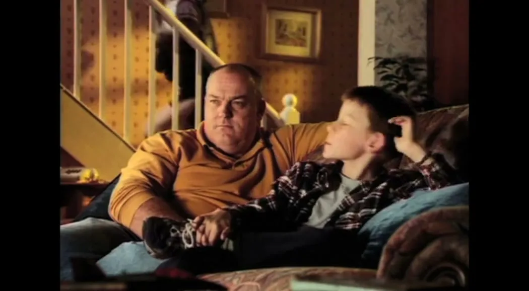 A father and son sit on a couch in their living room, watching TV, as another person walks up the stairs in the background.