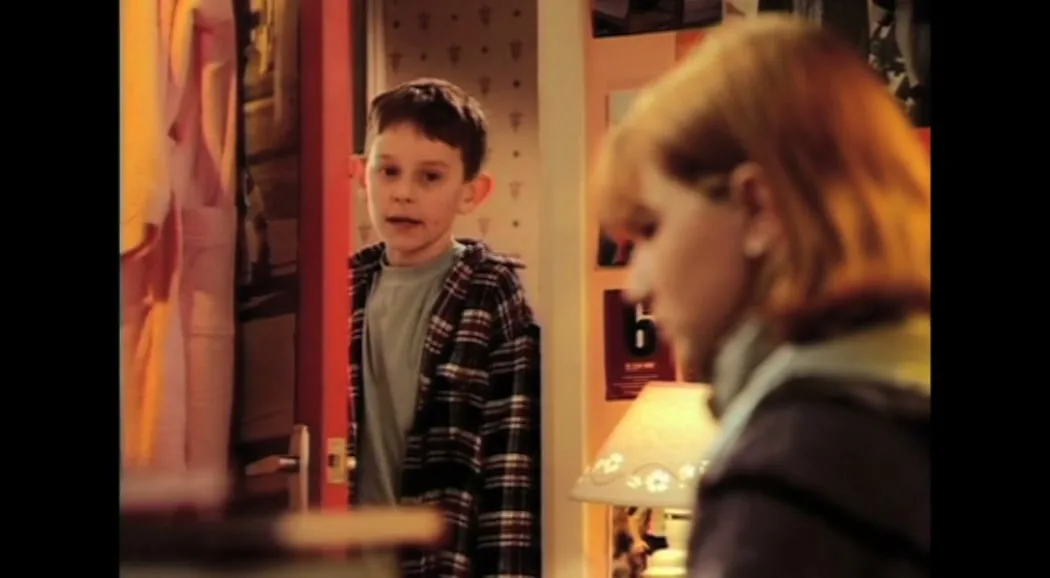 A young girl with red hair looks down sadly as her younger brother, a boy with brown hair, enters the room and looks at her with concern.