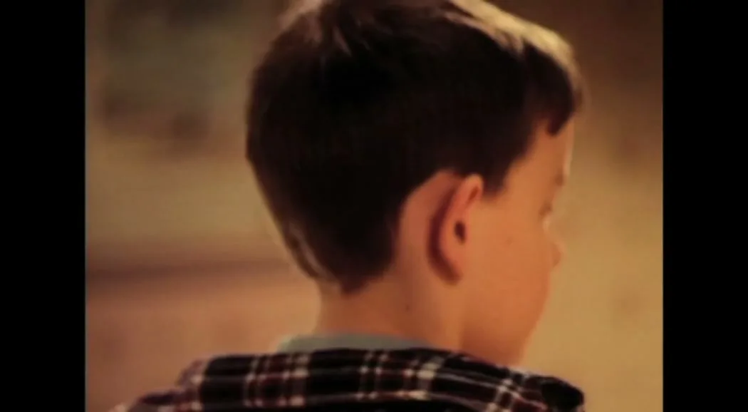 A young boy with big ears and a plaid shirt looks surprised, then turns his head and walks away from the camera in a home interior.