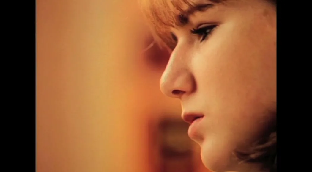 A young woman with reddish-brown hair and light makeup is shown in profile, looking down with a neutral expression in a warm-toned interior.