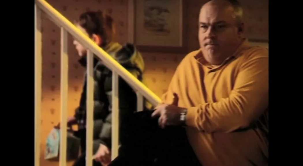 A man in an orange shirt and a boy in a black jacket are seen on a staircase in a house, with the man pulling the boy up the stairs.