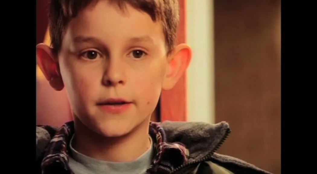 A young boy with brown hair and a grey hooded jacket looks directly at the camera, his expression shifting slightly, in an interior setting.