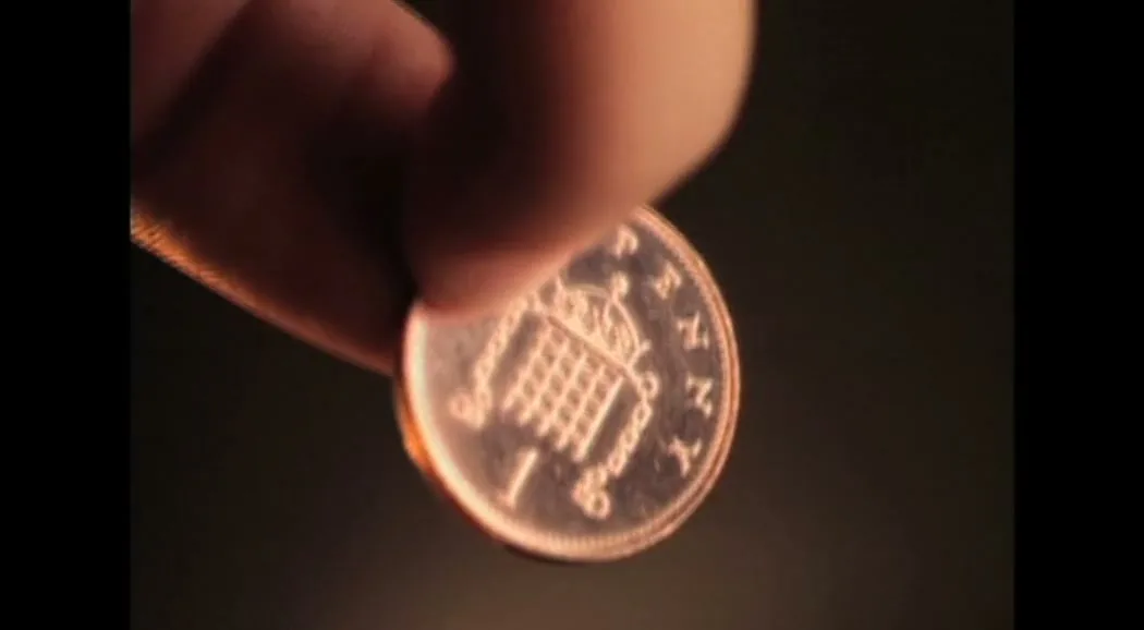A close-up shot shows a person's fingers holding a shiny copper coin, with a dark, blurry background.