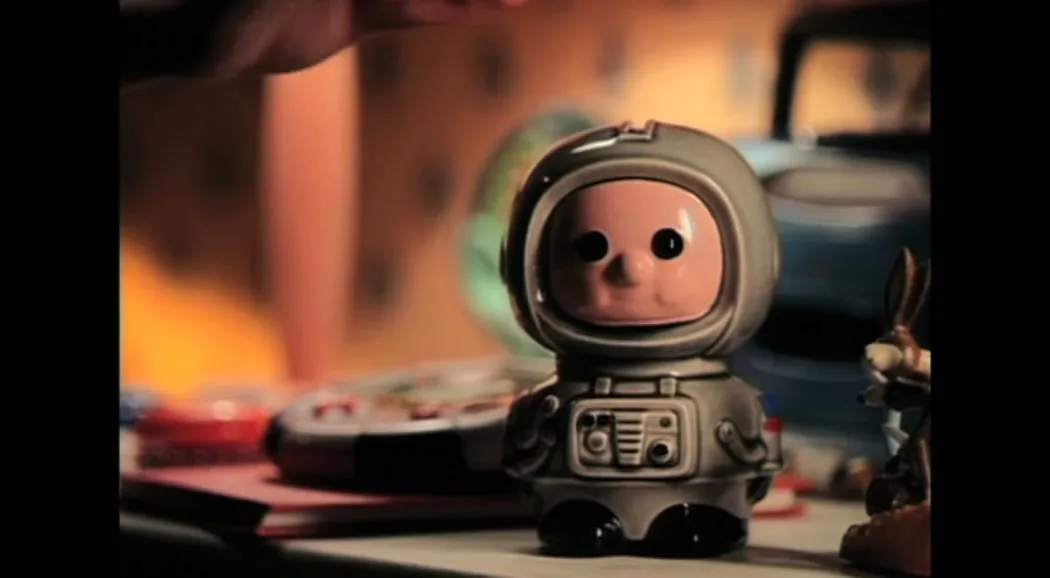 A child's hand drops a coin into a grey astronaut-shaped piggy bank with a pink face, on a cluttered desk in a bedroom.