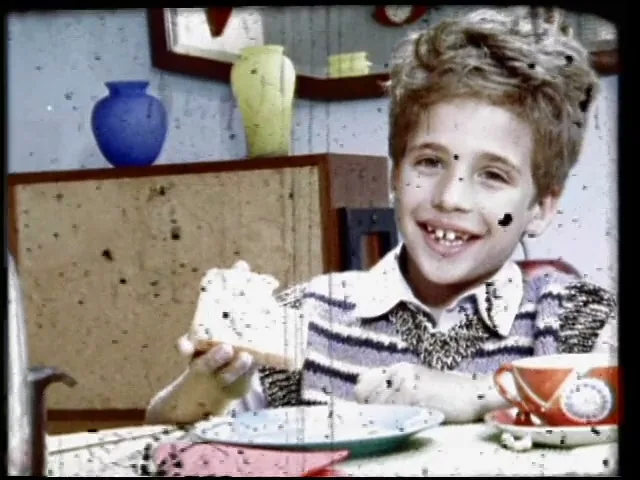 A young boy with curly hair, wearing a striped sweater, smiles at the camera while holding a piece of bread at a table in a colorful room.