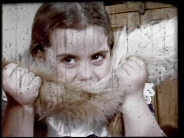A young girl with brown hair and a white collar plays with a fluffy, light brown object, holding it up to her face in an interior setting.