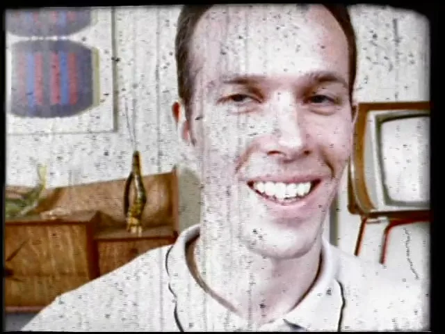 A man with short brown hair and a white polo shirt smiles at the camera, as static and film grain distort the image in a retro living room.