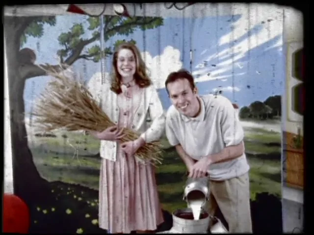 A smiling man and woman stand in front of a painted backdrop, the woman holding wheat and the man pouring milk into a churn.